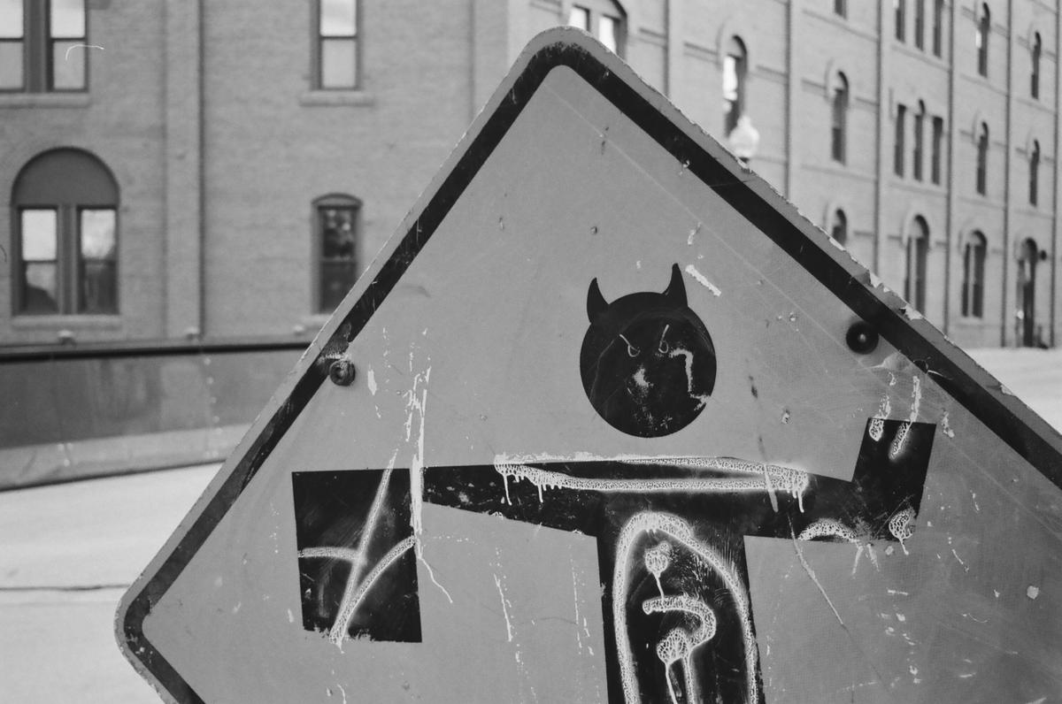 A close-up of a triangular street sign with graffiti on it is shown in black and white.