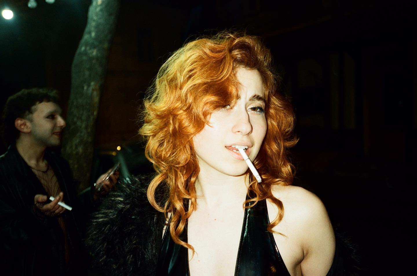 A woman with red hair and a shiny black top holds a cigarette in her mouth while a man in dark clothing stands behind her.