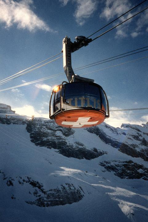 A cable car with a Swiss flag on its underside is suspended over a snow-covered mountain landscape under a blue sky with clouds.