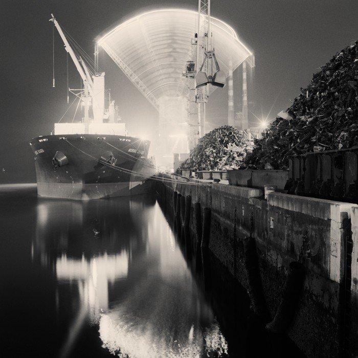 A large ship is docked next to a pier with piles of scrap metal under a brightly lit arched structure at night.