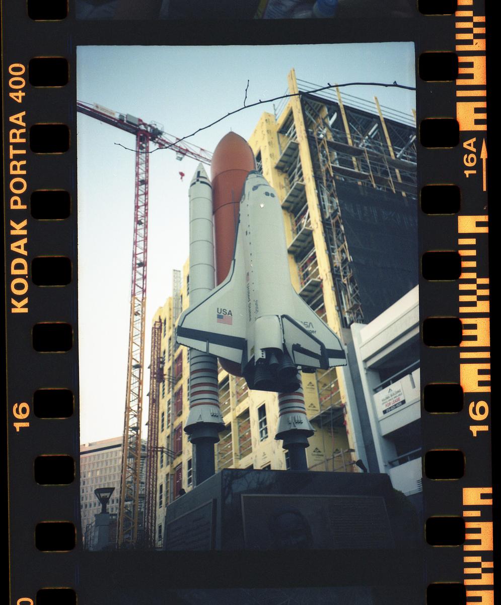 A model of a space shuttle stands on a pedestal in front of a building under construction with a crane.