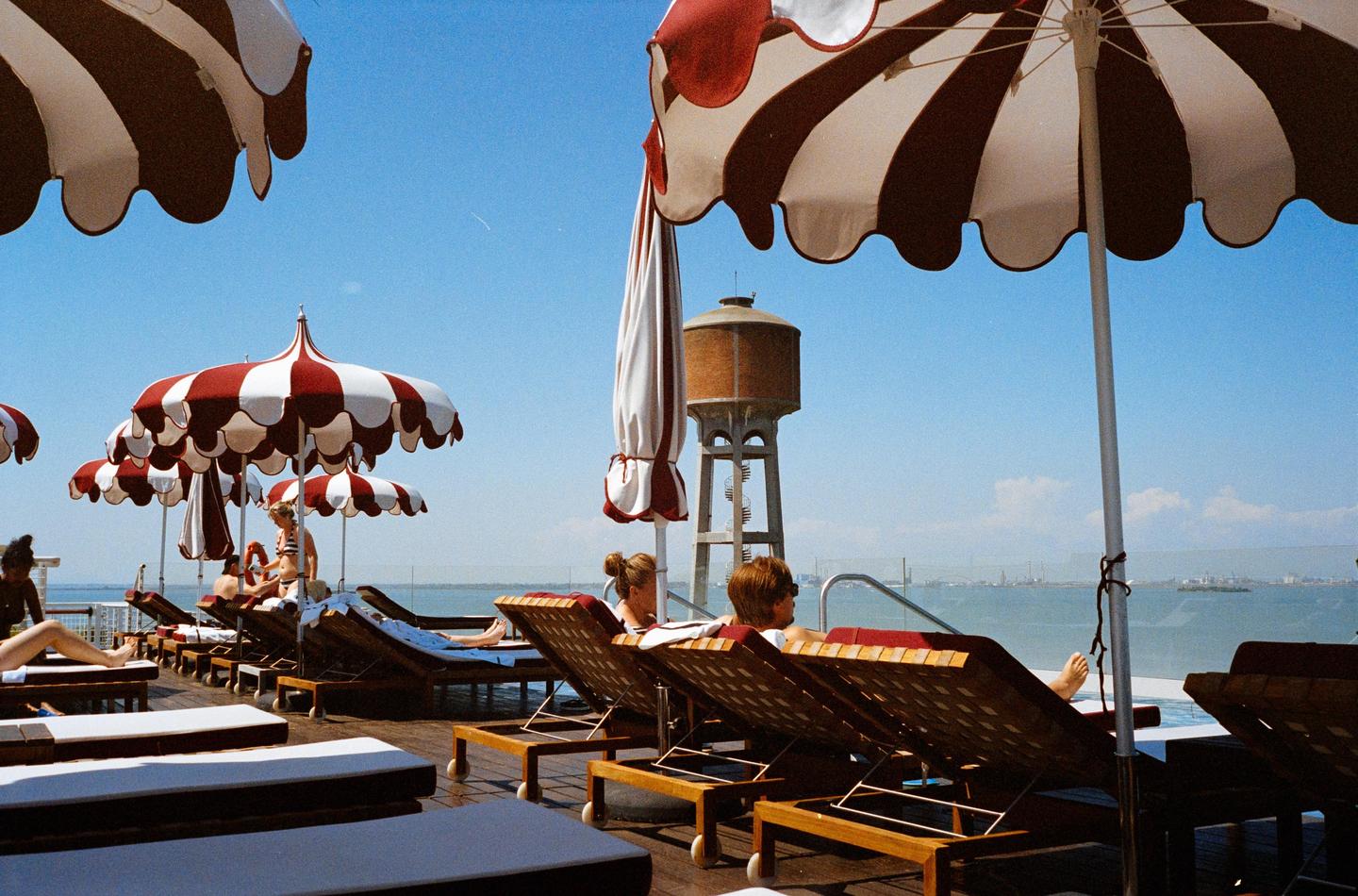 People relax on lounge chairs under striped umbrellas on a sunny deck overlooking a body of water with a water tower in the distance.