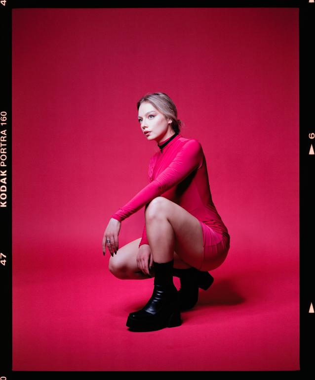A woman in a pink dress and black boots crouches against a solid pink background.