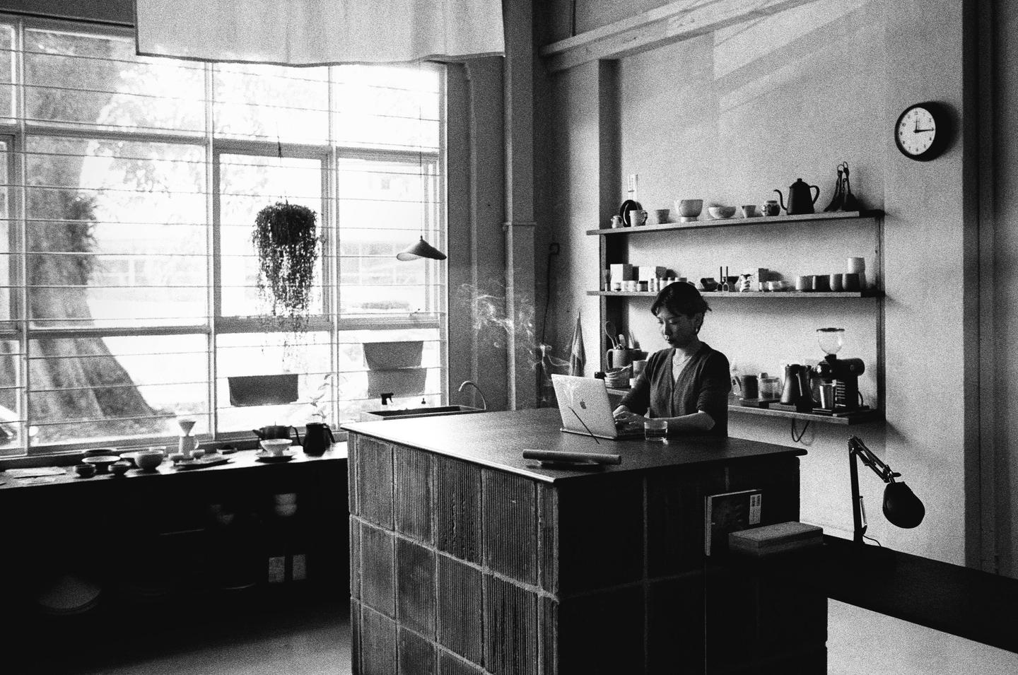 A woman sits at a counter with a laptop and a glass of water, with shelves of pottery and a large window in the background.