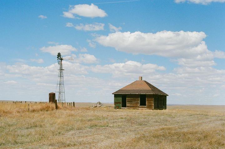 An abandoned wooden house with a windmill stands in a vast field under a blue sky with clouds.