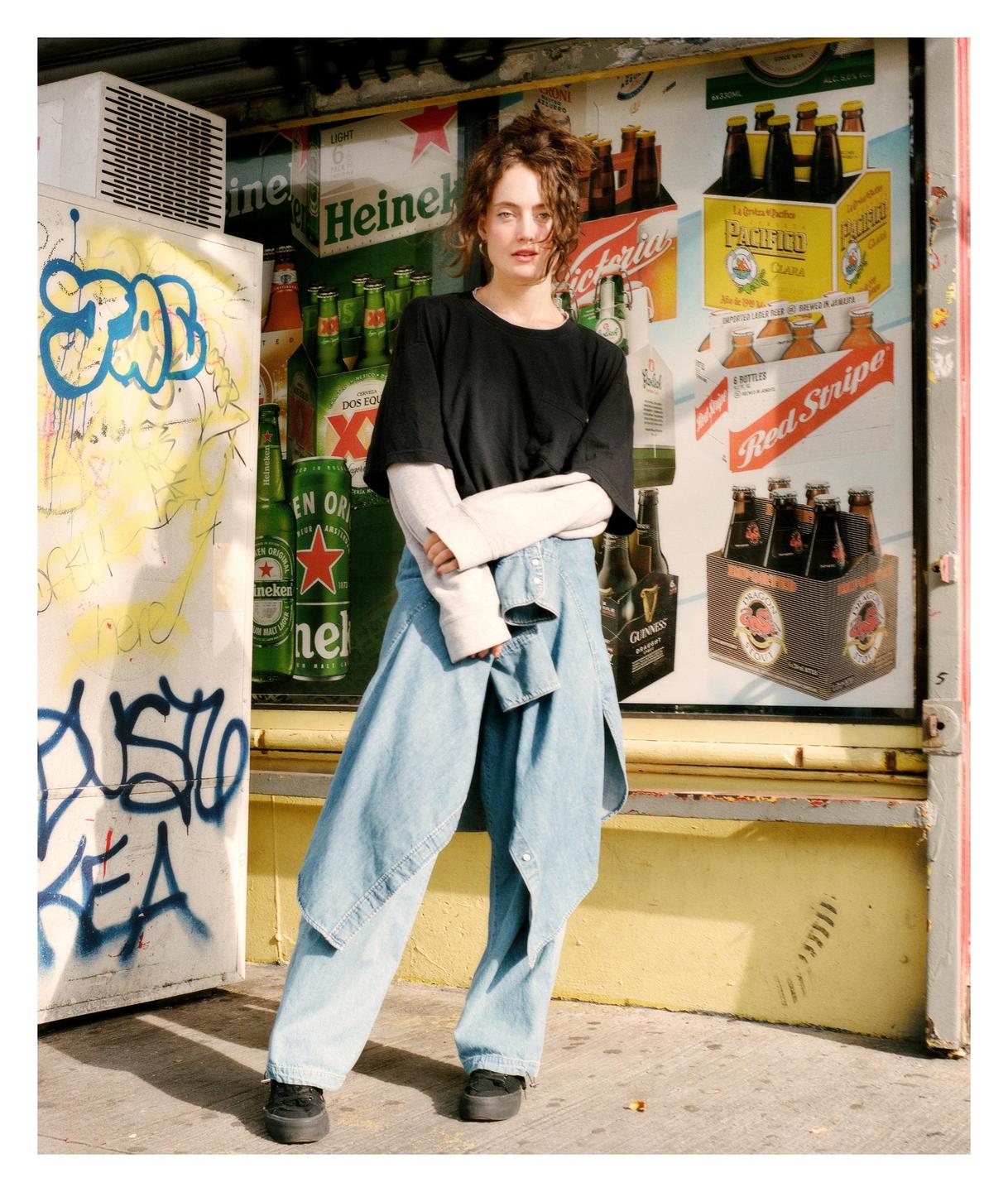 A woman stands in front of a storefront with beer advertisements and graffiti.