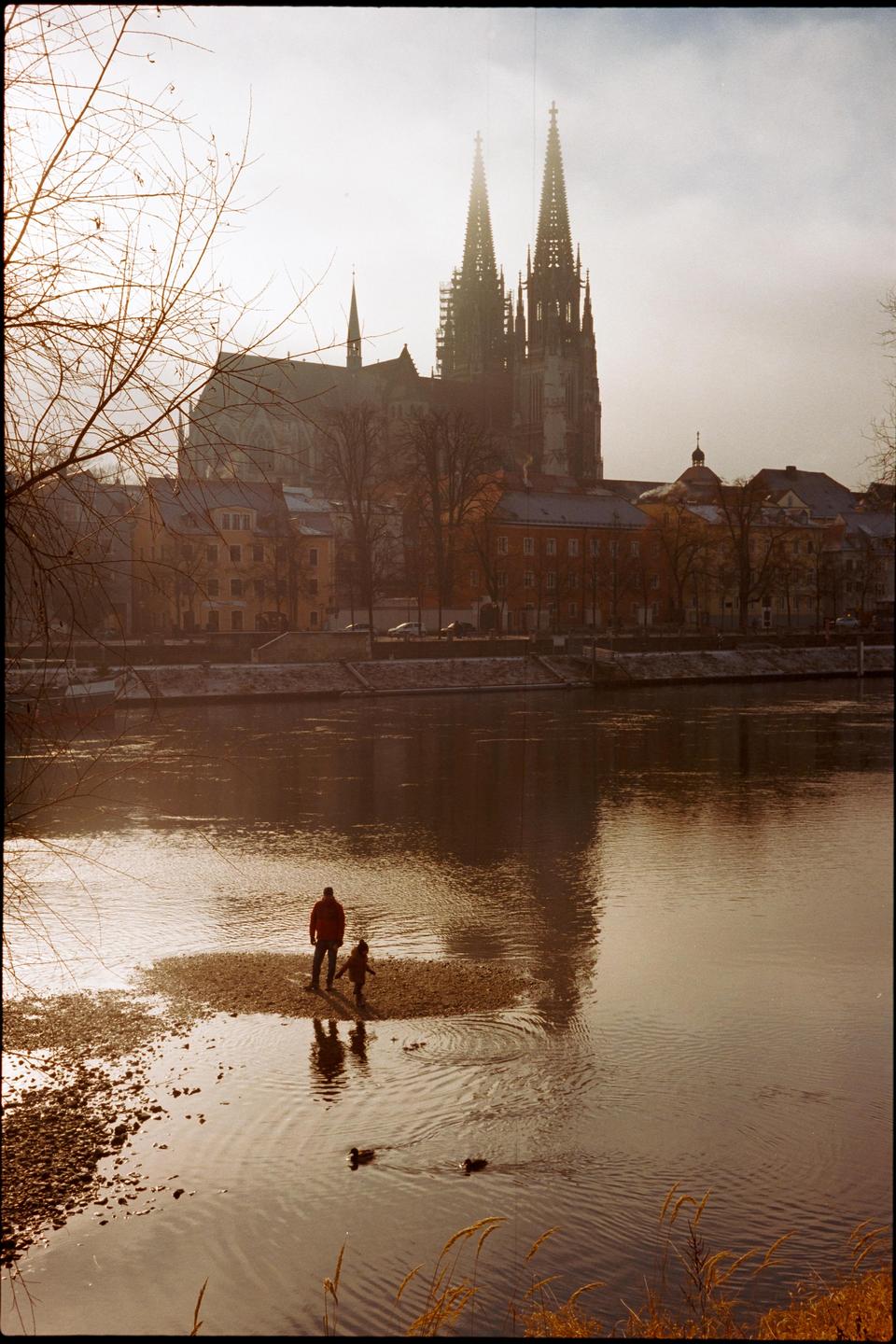 A man and a child stand on a small island in a river with a city and cathedral in the background.