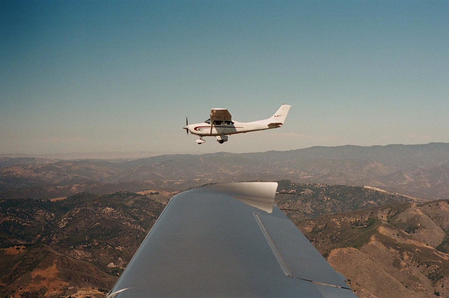 A small white airplane flies above a mountainous landscape, with the wing of another aircraft visible in the foreground.