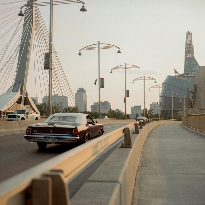 A brown convertible drives on a bridge with a modern building in the background.