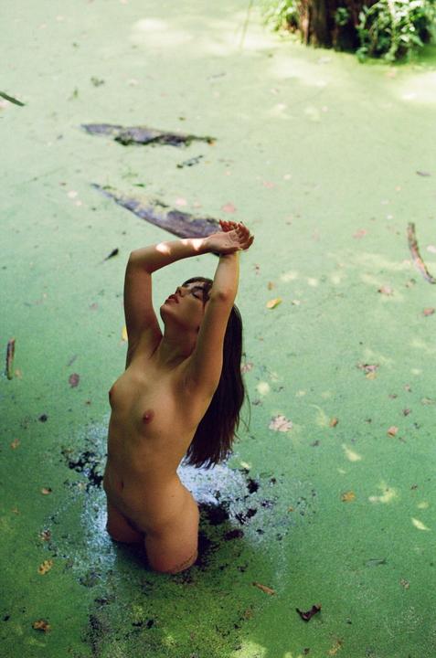 A woman stands in a body of water covered in green algae, with her arms raised above her head.