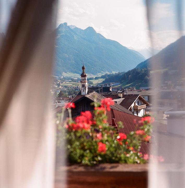 A view of a village with a church steeple and mountains in the background, framed by curtains and red flowers.