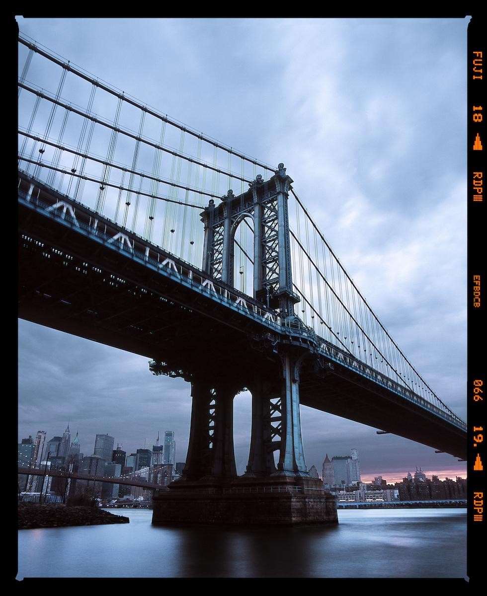 A large suspension bridge spans a body of water with a city skyline in the background under a cloudy sky.