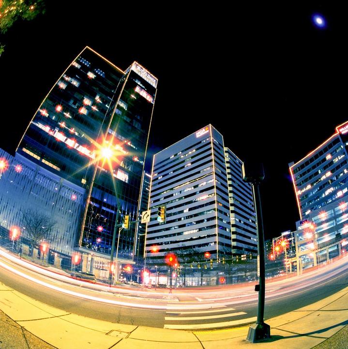 A fisheye view of a city street at night shows tall buildings illuminated by streetlights and light trails from moving vehicles.