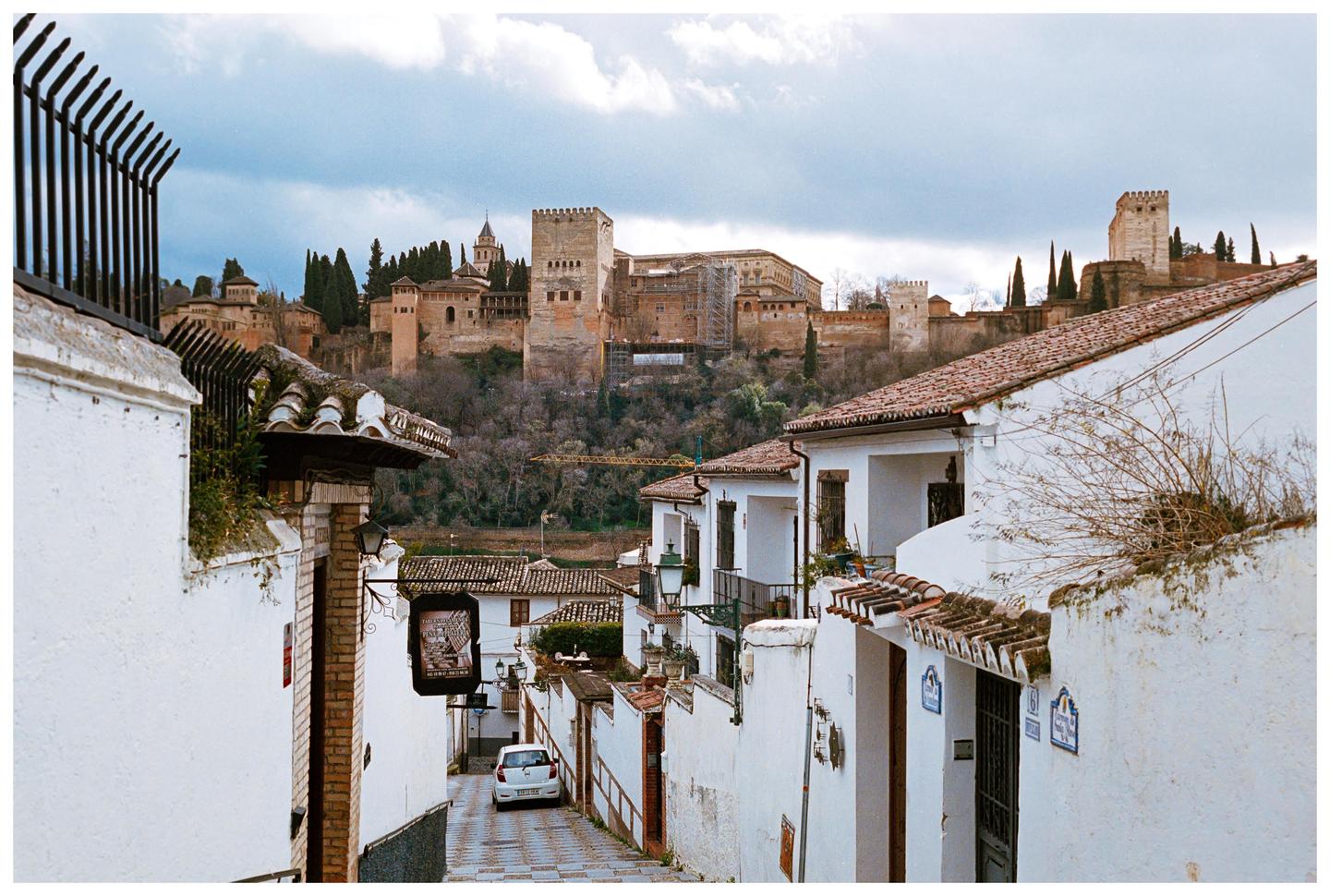 A narrow street with white buildings and terracotta roofs leads down to a small white car, with a large castle on a hill in the background.