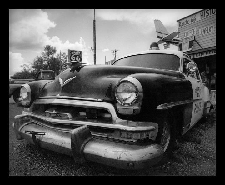 A vintage police car is parked on a gravel lot with a "Route 66" sign and a building with "Coffee" and "Jewelry" signs in the background.