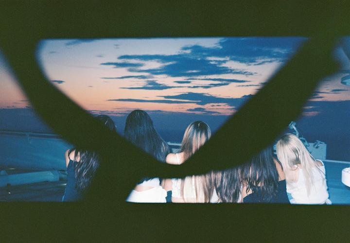 Four women with long hair are seen from behind, looking out at a sunset over the ocean.