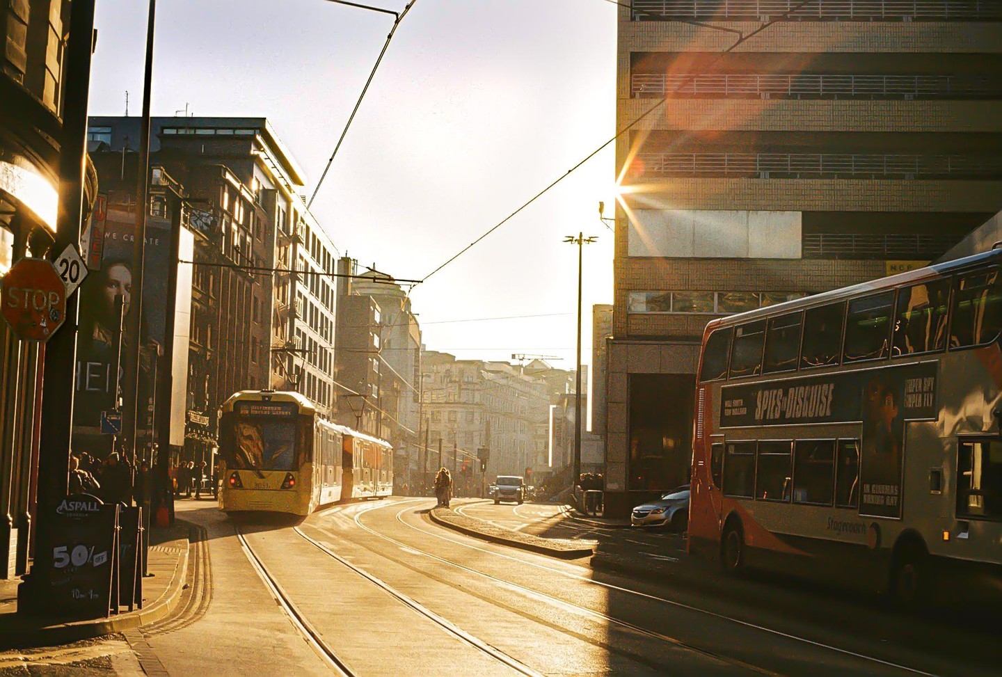 A double-decker bus and a tram are on a city street at golden hour, with sunlight flaring from behind a building.