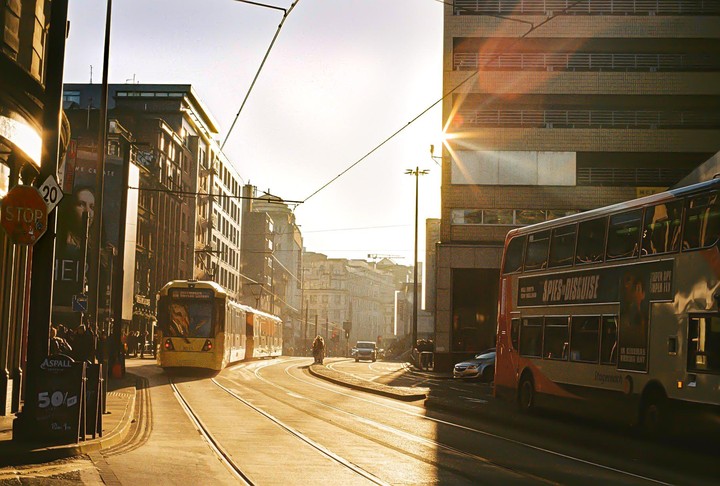 A double-decker bus and a tram are on a city street at golden hour, with sunlight flaring from behind a building.