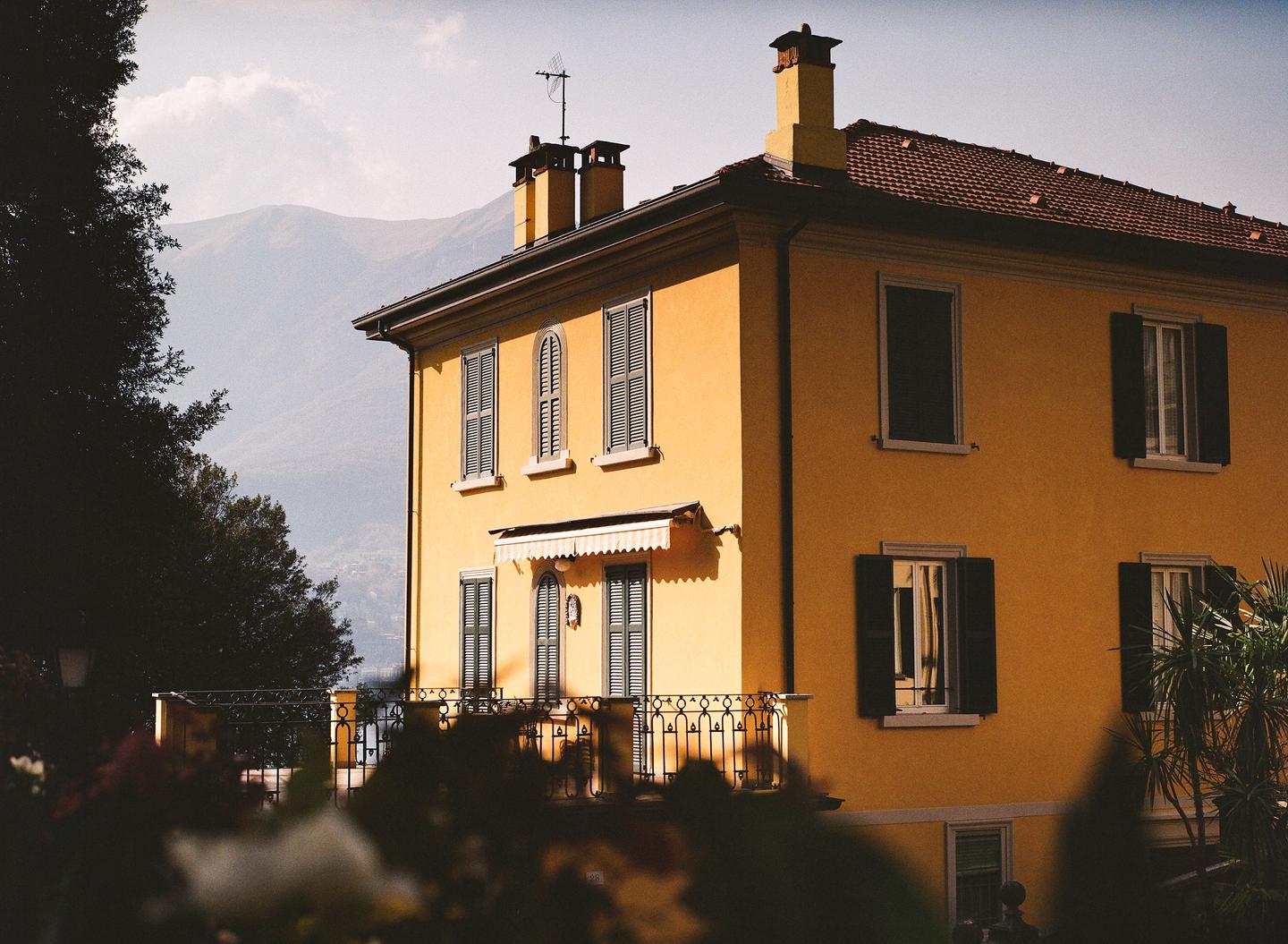 A golden building with a red tile roof and many windows with shutters stands in front of a hazy mountain.