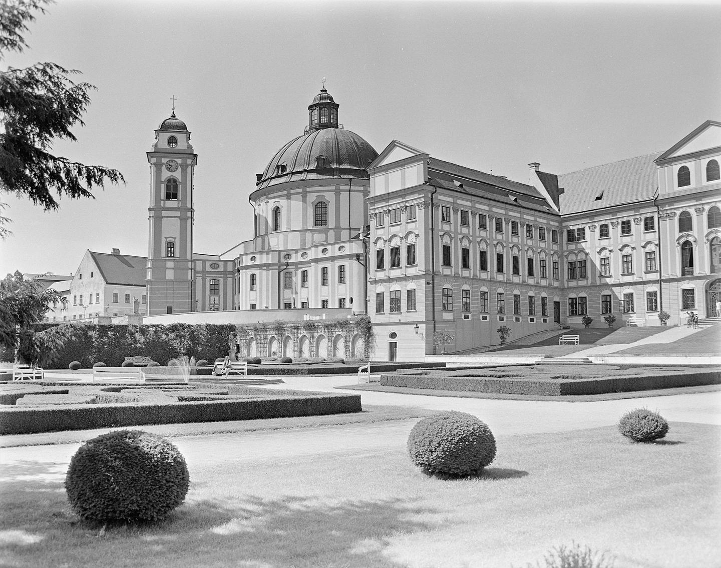 A large baroque building with a domed roof and a clock tower overlooks a formal garden with manicured hedges and spherical bushes.