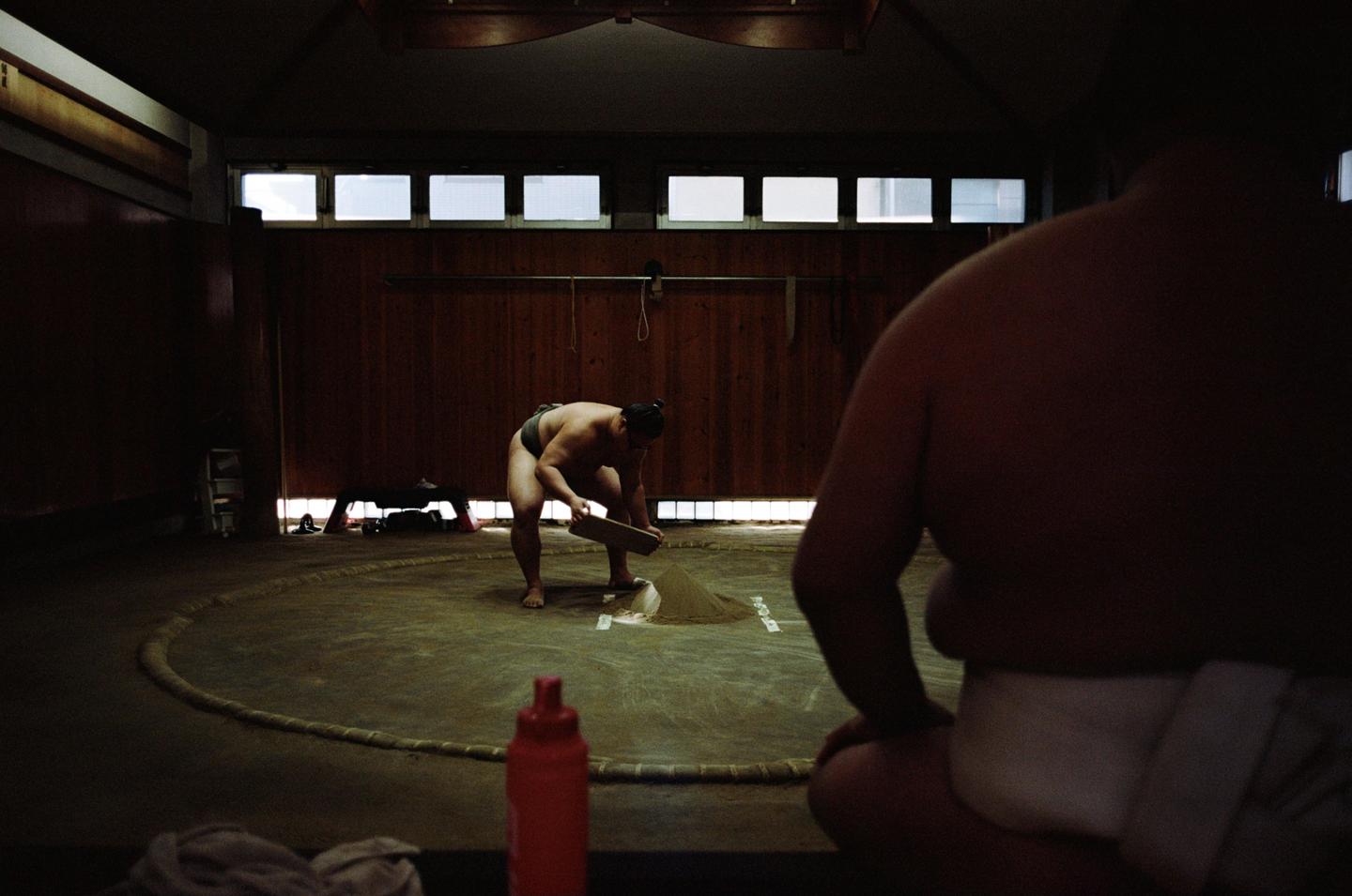 A sumo wrestler prepares the ring while another sumo wrestler sits in the foreground.