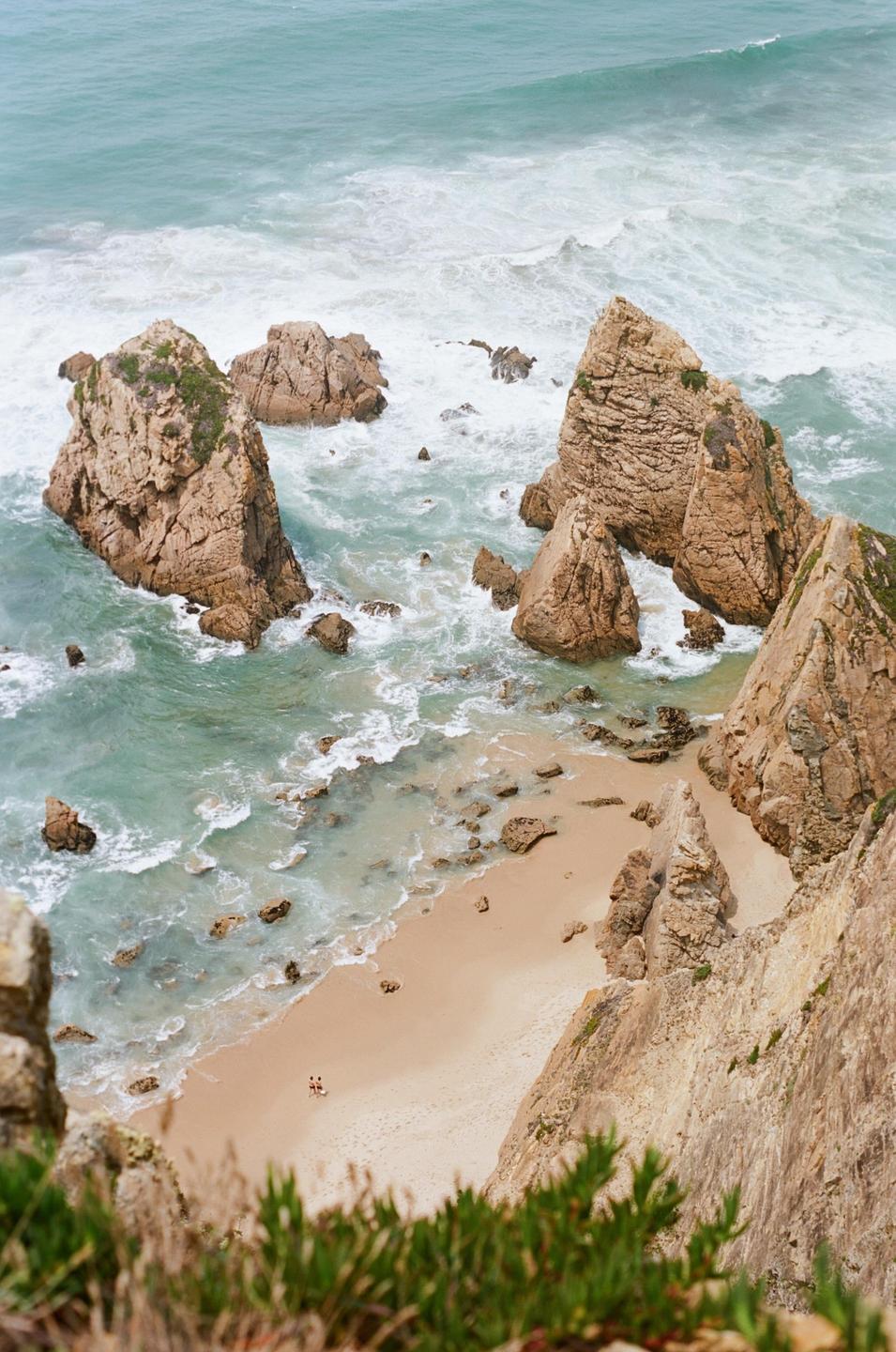 A high-angle view shows a sandy beach with two people, surrounded by large rock formations and ocean waves.