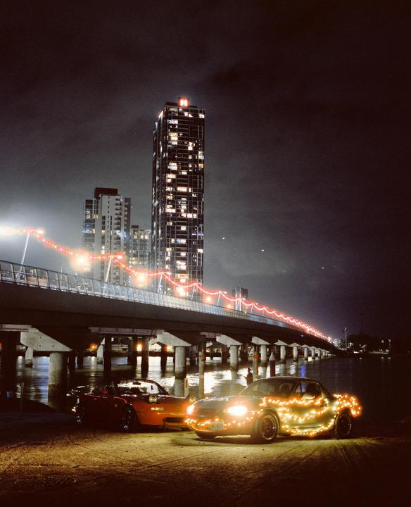Two cars decorated with string lights are parked under a bridge at night, with a tall building in the background.
