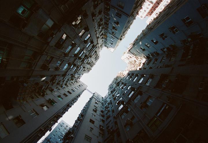 Looking up from a narrow courtyard, tall apartment buildings with many windows and air conditioning units rise towards a bright sky.