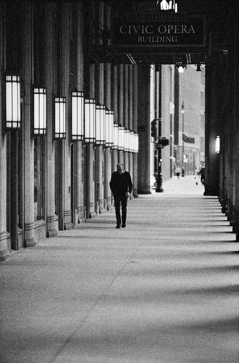 A man walks down a long, columned walkway with many illuminated lamps on the left and a sign for the Civic Opera Building overhead.