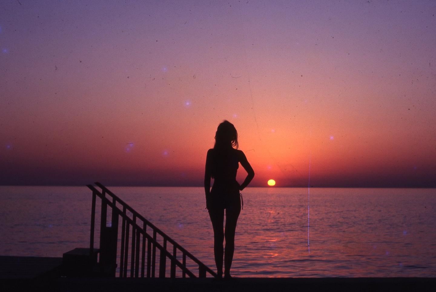 A woman stands in silhouette on a dock overlooking the ocean at sunset.