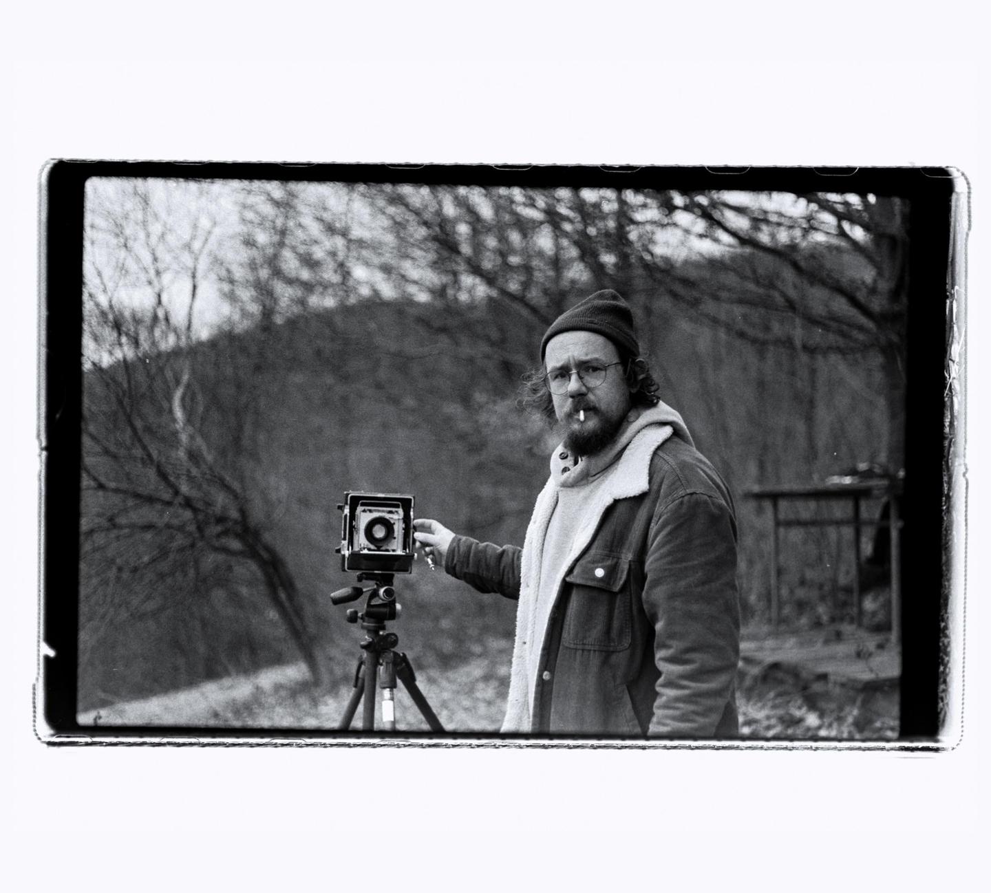 A man with a cigarette in his mouth stands next to a camera on a tripod in a wooded area.