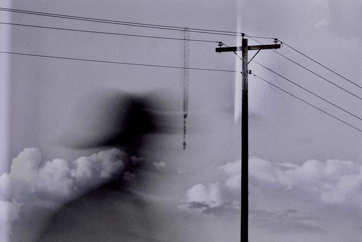 A telephone pole with wires stands against a cloudy sky with a faint, dark, ghost-like figure on the left.