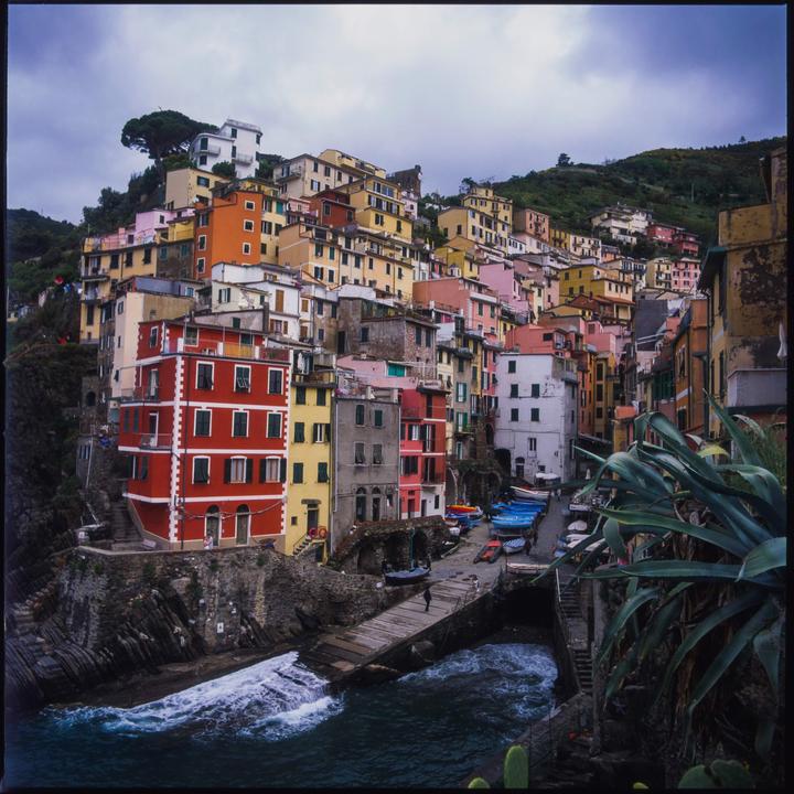 A colorful village with houses built into a hillside overlooks a small harbor with boats and crashing waves.