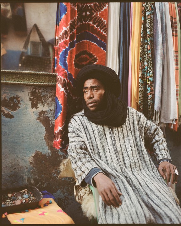 A man wearing a black turban and a striped robe sits in front of colorful fabrics.