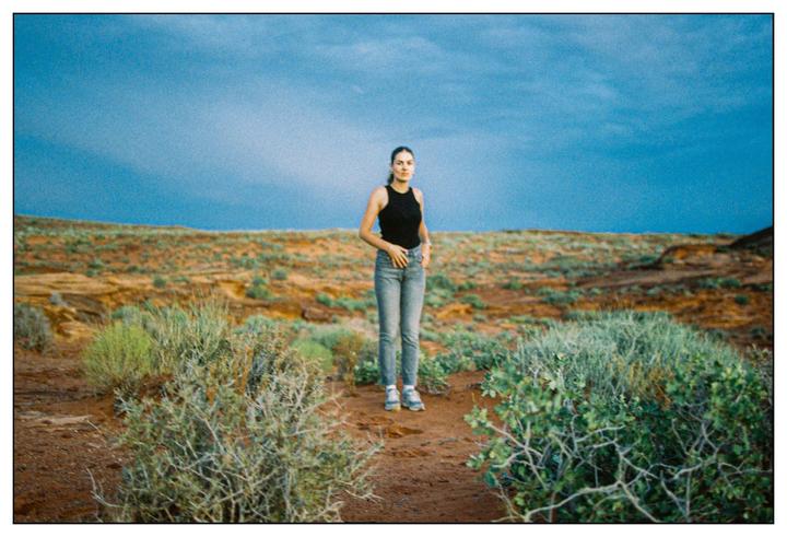 A woman stands in a desert landscape with sparse vegetation under a blue sky.