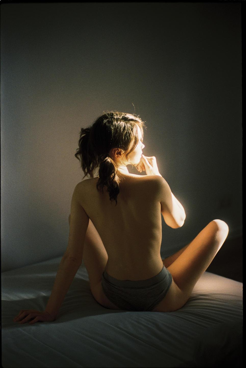 A woman with a ponytail sits on a bed with her back to the viewer, looking over her shoulder.