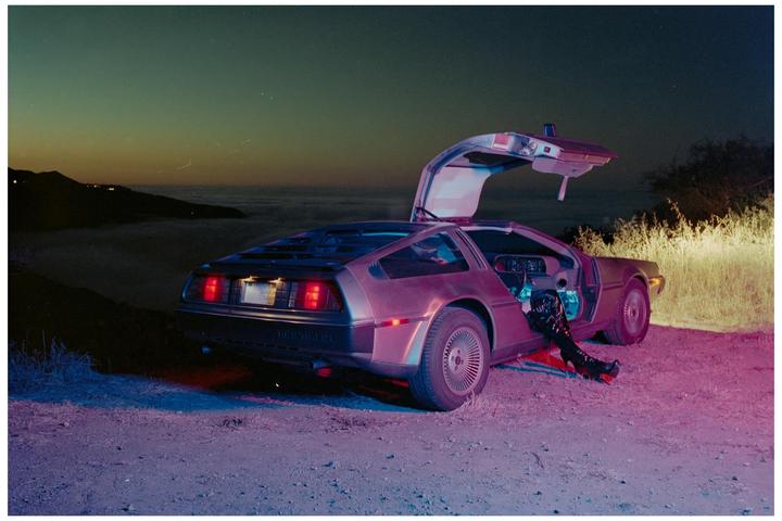 A DeLorean with its gull-wing door open is parked on a dirt road overlooking a cloud-filled valley at dusk.