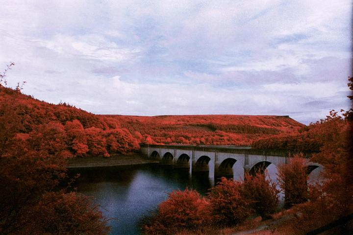 A bridge with multiple arches spans a body of water, surrounded by hills covered in red foliage under a cloudy sky.
