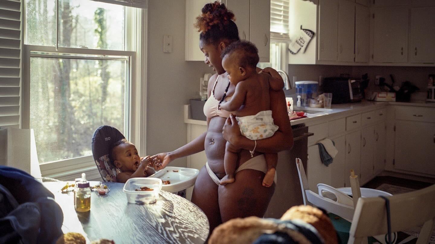 A woman holds one baby while another baby sits in a high chair at a table in a kitchen.