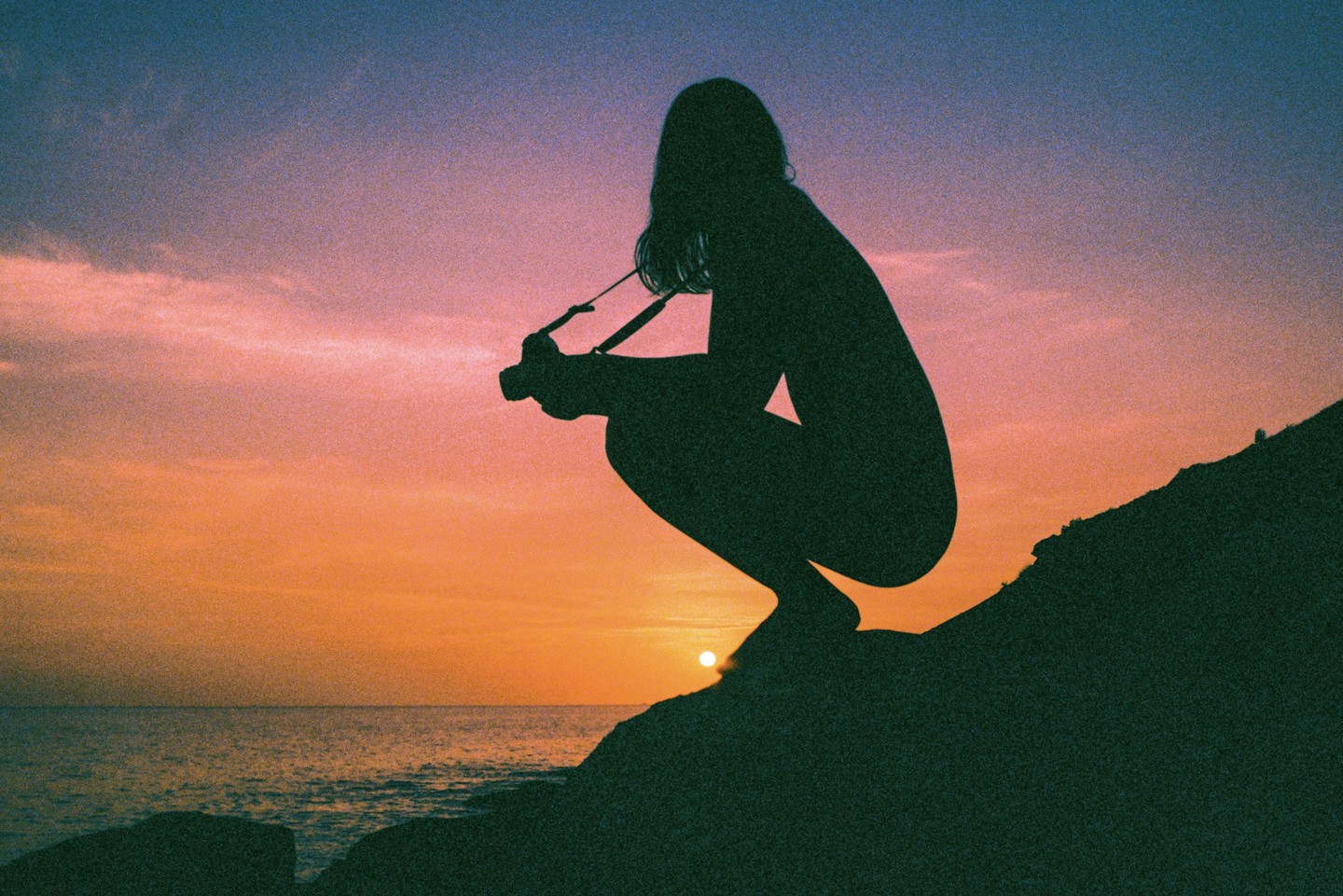 A woman crouches on a rocky outcrop by the sea, silhouetted against a vibrant sunset.