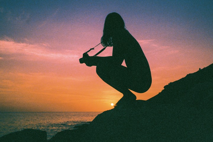 A woman crouches on a rocky outcrop by the sea, silhouetted against a vibrant sunset.
