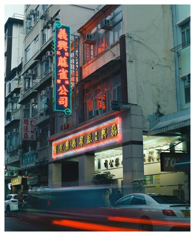 A street in Kowloon City is illuminated by neon signs and the light trails of passing vehicles at night.