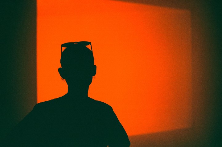 A person in silhouette with sunglasses on their head stands against an orange wall.