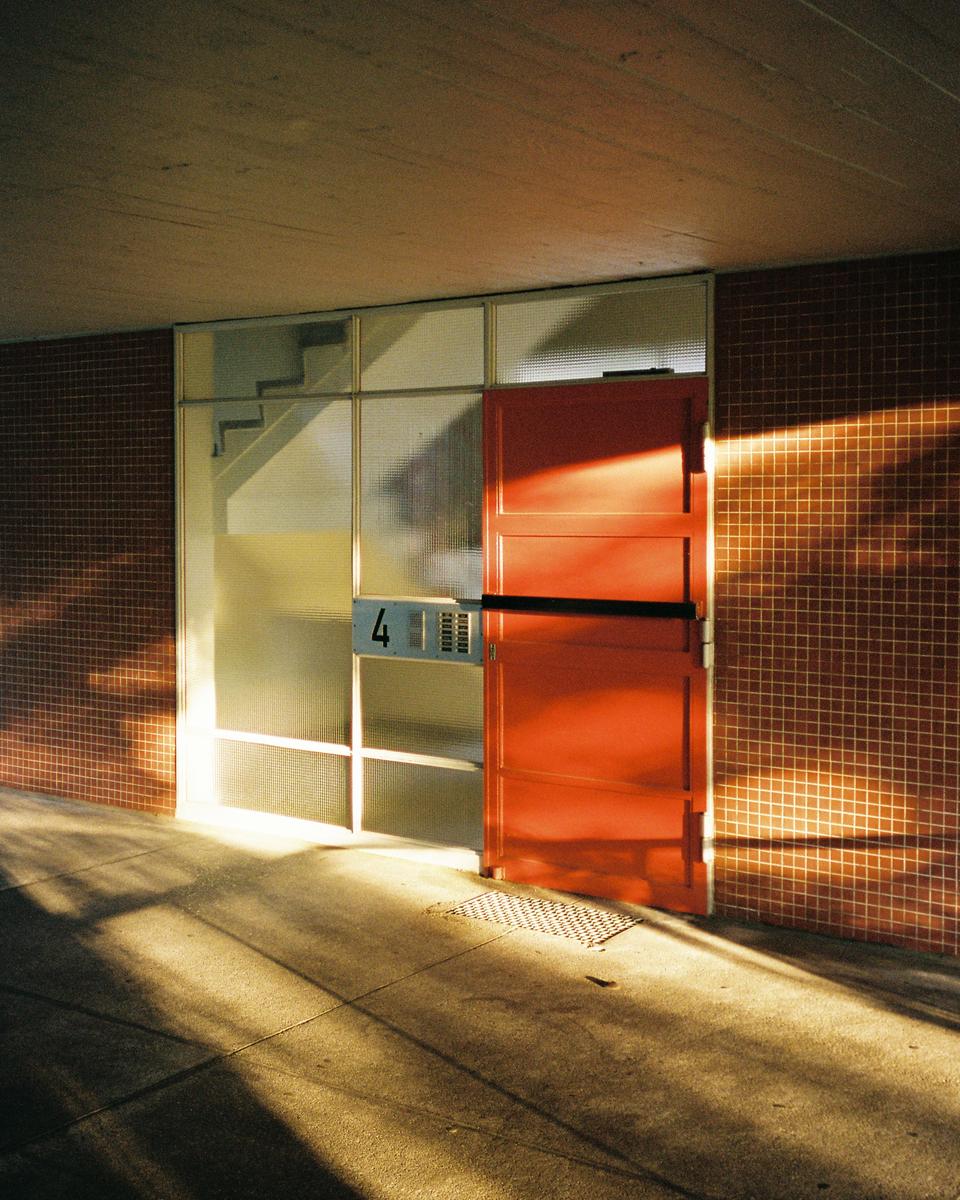 Sunlight casts shadows on a red door and a tiled wall next to a window with the number four.