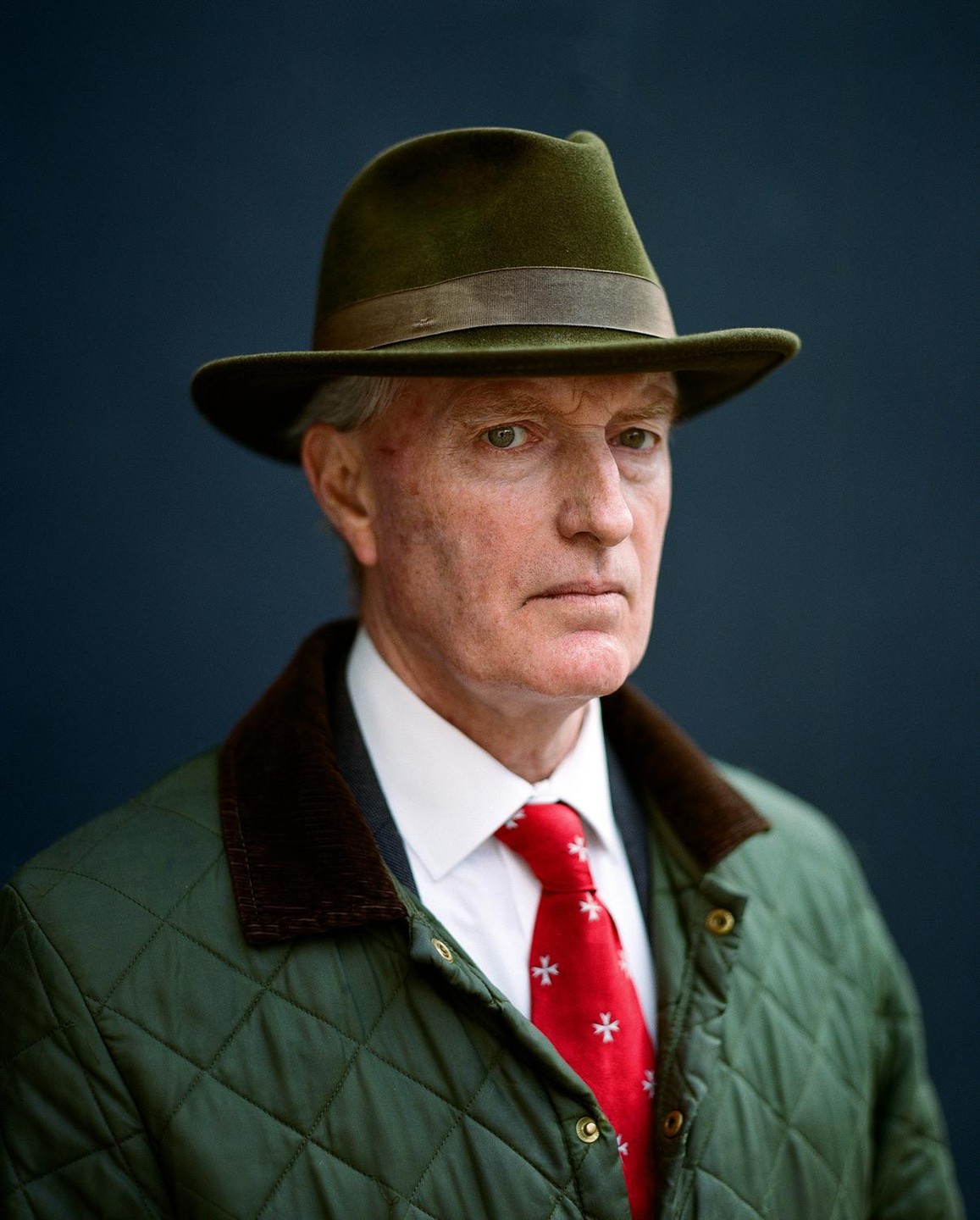 A man wearing a green hat, a green quilted jacket, and a red tie with white crosses is looking directly forward.