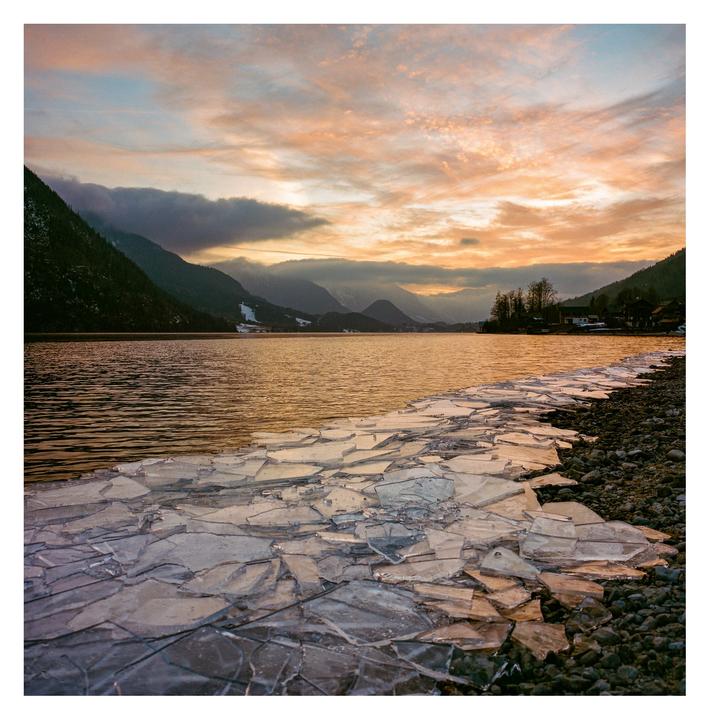 A lake with drift ice on its shore is surrounded by mountains under a colorful sunset sky.
