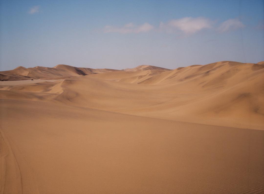 A vast desert landscape features rolling sand dunes under a clear sky with a few clouds.