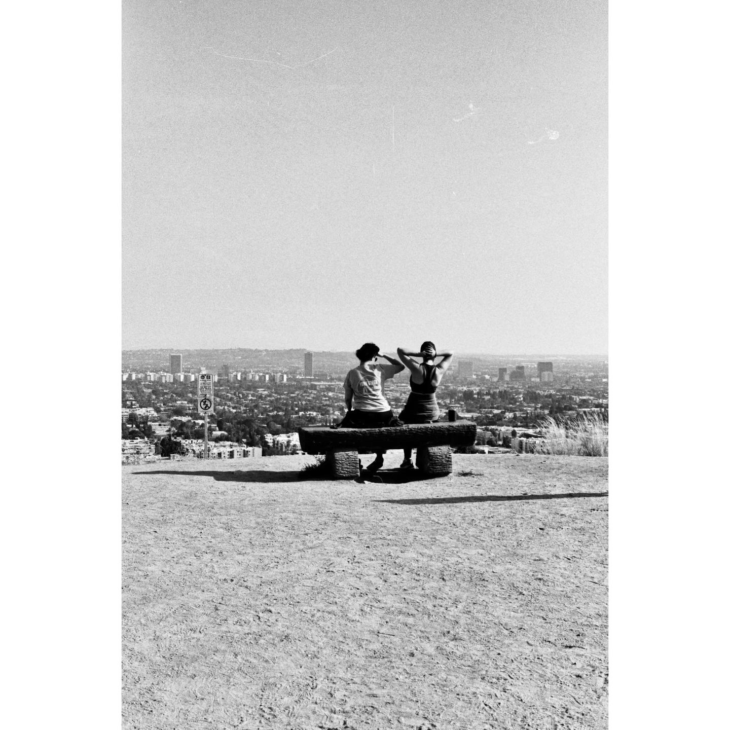 Two people sit on a wooden bench overlooking a city.