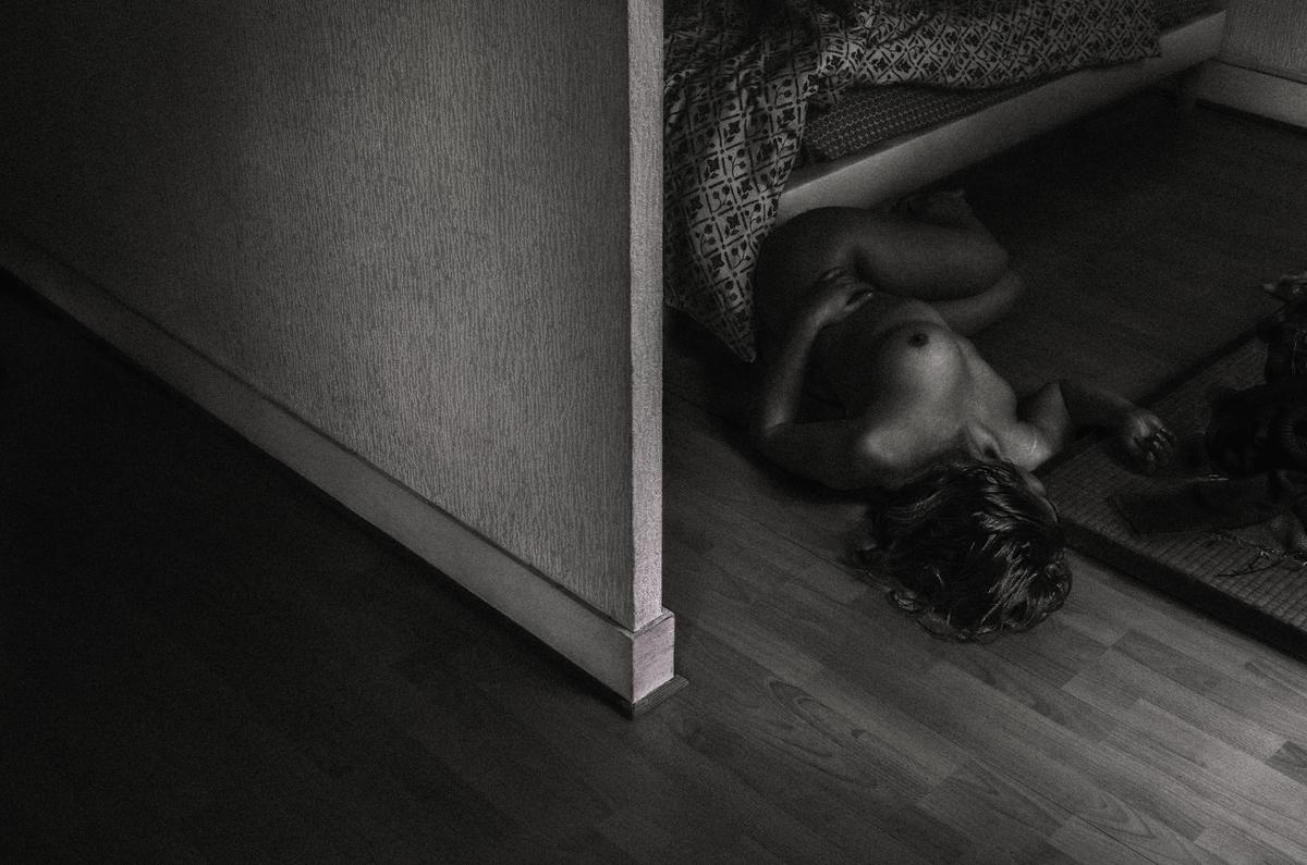 A nude woman lies on a wooden floor next to a wall.