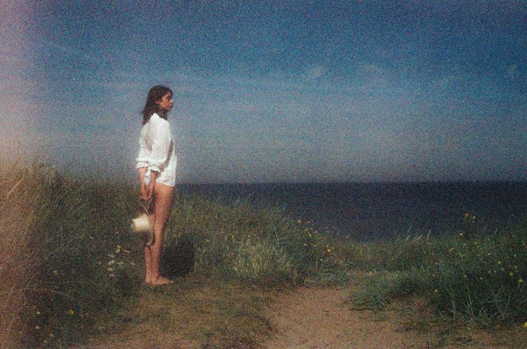 A woman stands barefoot in tall grass on a dune overlooking the ocean.